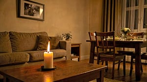 A cozy living room with a candle burning on a coffee table, a photo frame on the wall, and a dining table set for a meal with an empty chair, under soft, warm lighting.
