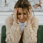 A woman sitting on a sofa holding her head, expressing stress and frustration indoors.