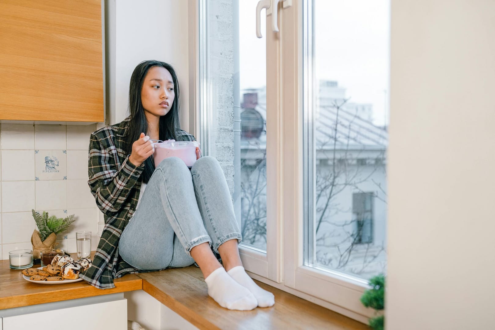 An Asian woman sits by the window, enjoying dessert with an introspective look.