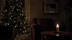 A dimly lit living room with a Christmas tree, a single candle burning on a table, and a photo frame visible in the background, symbolizing the presence of memories and the absence of a loved one during the holidays.