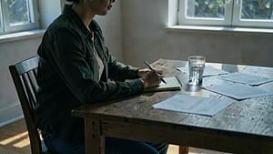 A person sitting at a table with a notebook, pen, and glass of water, in a quiet, well-lit room with natural light coming through a window.