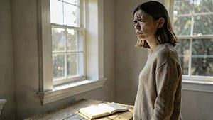 A person standing in a quiet, sunlit room with a journal on a wooden table nearby, looking out the window in a calm and reflective manner.