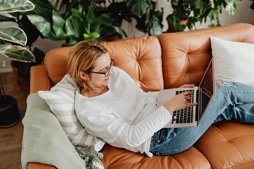pexels-photo-5903077-5903077 Woman in casual attire using a laptop on a couch surrounded by plants, emphasizing remote work culture.