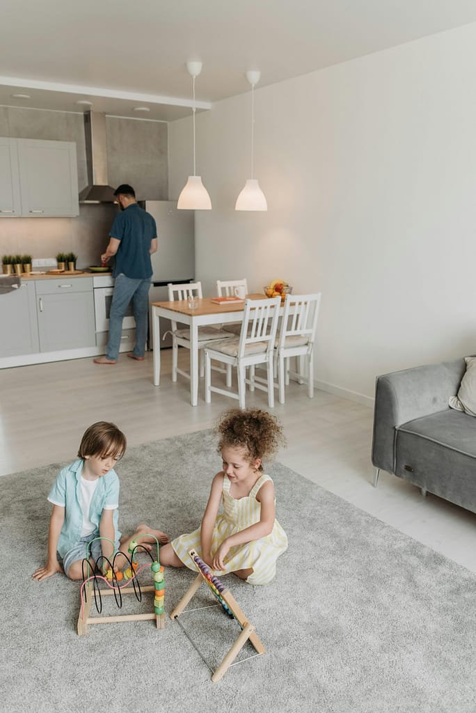 pexels-photo-8763143-8763143 Two children playing on carpet with wooden toys while adult cooks in modern kitchen.