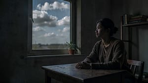 A person sitting at a table in a quiet room, looking out the window with a calm expression. A plant is visible on the windowsill, and there are books on a nearby shelf.