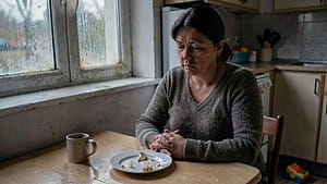 A woman sits at a kitchen table, staring blankly at an uneaten meal. Natural light enters through a window, and a toy lies on the floor nearby.