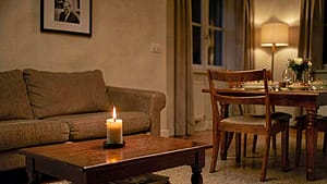 A cozy living room with a candle burning on a coffee table, a photo frame on the wall, and a dining table set for a meal with an empty chair, under soft, warm lighting.