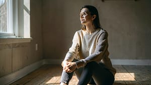 A person sitting in a quiet, sunlit room, hands resting on their knees, surrounded by soft natural light. The room is simple and calm.